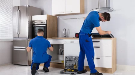 Zwei Küchenbauer in blauer Arbeitskleidung montieren eine neue Küche. Einer kniet vor einem offenen Unterschrank und arbeitet daran, während der andere sich über eine Arbeitsplatte beugt und ein Gerät installiert. Auf dem Boden steht ein offener Werkzeugkasten. Die Küche ist modern eingerichtet, mit einem großen Edelstahlkühlschrank und weißen Schränken.
