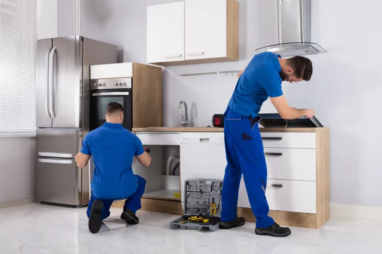 Zwei Küchenbauer in blauer Arbeitskleidung montieren eine neue Küche. Einer kniet vor einem offenen Unterschrank und arbeitet daran, während der andere sich über eine Arbeitsplatte beugt und ein Gerät installiert. Auf dem Boden steht ein offener Werkzeugkasten. Die Küche ist modern eingerichtet, mit einem großen Edelstahlkühlschrank und weißen Schränken.