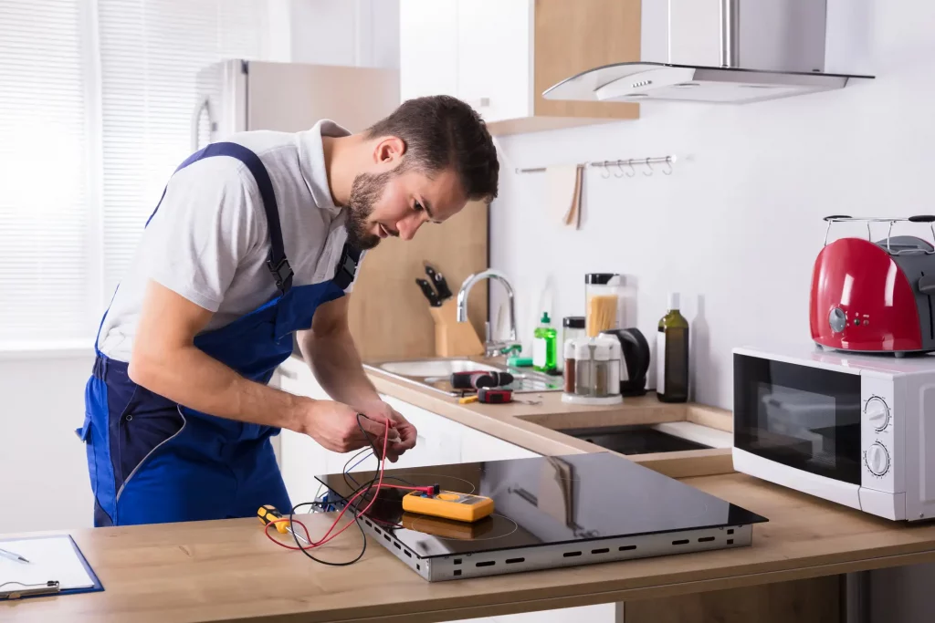 Ein Küchenbauer in blauer Arbeitskleidung installiert ein Kochfeld in einer modernen Küche. Er überprüft die Verkabelung mit einem Multimeter. Auf der Arbeitsplatte stehen verschiedene Küchenutensilien. Im Hintergrund sind ein Edelstahlkühlschrank, eine Spüle und weiße Schränke zu sehen.