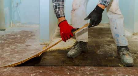 Ein Handwerker in weißer, mit Farbflecken übersäter Hose, einem schwarz-weiß karierten Hemd und roten sowie schwarzen Handschuhen entfernt alten Laminat in einem Raum. Der Boden darunter ist dunkel und abgenutzt. Die Wände sind hellblau gestrichen.