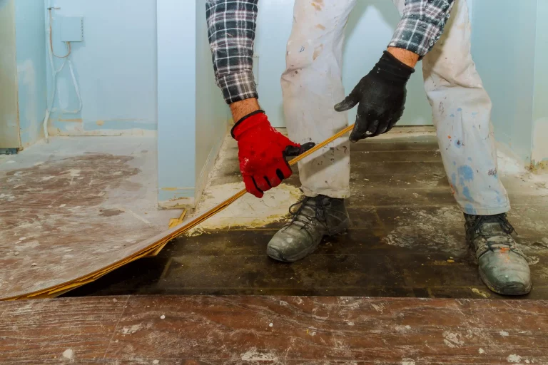 Ein Handwerker in weißer, mit Farbflecken übersäter Hose, einem schwarz-weiß karierten Hemd und roten sowie schwarzen Handschuhen entfernt alten Laminat in einem Raum. Der Boden darunter ist dunkel und abgenutzt. Die Wände sind hellblau gestrichen.