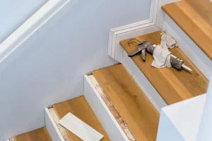 Eine Treppe wird mit Laminat verkleidet. Auf den Stufen liegen Werkzeuge, darunter eine Kartuschenpistole und Tücher. Teile der Treppe sind bereits mit Laminat verkleidet, während andere noch bearbeitet werden. Eine Leiste liegt auf einer der Stufen. Die Wände sind weiß gestrichen, und die Treppe hat weiße Seitenverkleidungen.