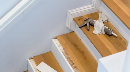 Eine Treppe wird mit Laminat verkleidet. Auf den Stufen liegen Werkzeuge, darunter eine Kartuschenpistole und Tücher. Teile der Treppe sind bereits mit Laminat verkleidet, während andere noch bearbeitet werden. Eine Leiste liegt auf einer der Stufen. Die Wände sind weiß gestrichen, und die Treppe hat weiße Seitenverkleidungen.
