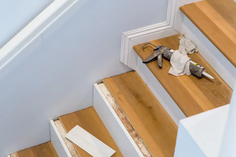 Eine Treppe wird mit Laminat verkleidet. Auf den Stufen liegen Werkzeuge, darunter eine Kartuschenpistole und Tücher. Teile der Treppe sind bereits mit Laminat verkleidet, während andere noch bearbeitet werden. Eine Leiste liegt auf einer der Stufen. Die Wände sind weiß gestrichen, und die Treppe hat weiße Seitenverkleidungen.