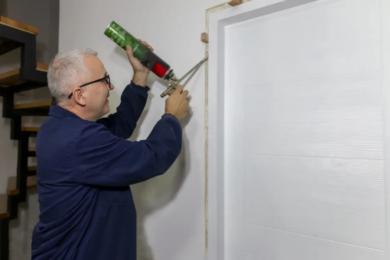 Ein Türenbauer mit grauen Haaren und Brille, der eine Türzarge einbaut. Er verwendet eine Schaum-Pistole, um die Türzarge in der Wand zu fixieren. Der Mann trägt eine dunkelblaue Arbeitsjacke. Im Hintergrund ist eine Holztreppe zu sehen. Das Bild zeigt den handwerklichen Prozess des Türzargeneinbaus im Innenbereich eines Hauses.