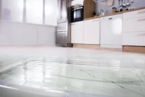 Eine moderne Küche mit weißer und hölzerner Einrichtung ist zu sehen, die von einem schweren Wasserschaden betroffen ist. Der Boden ist mit einer deutlichen Schicht Wasser bedeckt, wodurch kleine Wellen entstehen. Im Hintergrund befinden sich eine Edelstahlkühlschrank, ein Backofen, eine Spülmaschine und ein Waschbecken, die teilweise unscharf sind. An der Wand hängen Küchenutensilien. Das Licht, das durch die Fenster mit Jalousien einfällt, reflektiert auf der Wasseroberfläche. Die Szene zeigt eine deutliche Überflutung, die die gesamte Küche betrifft.