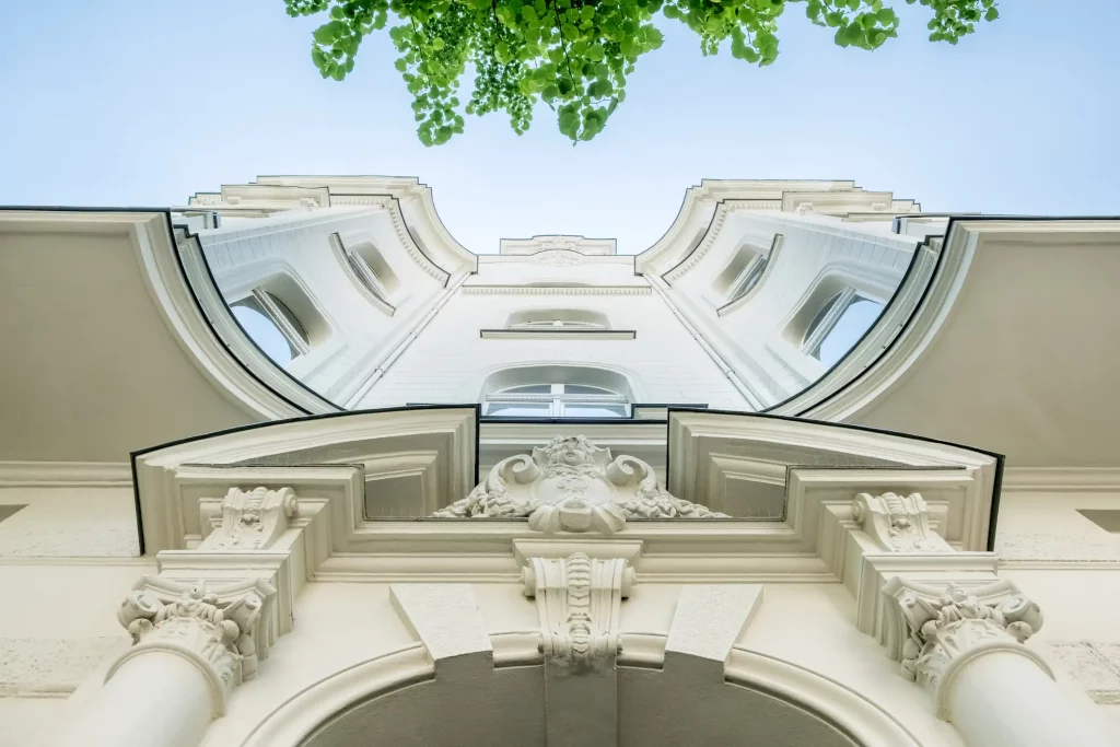 Blick von unten auf eine historische Fassade eines Gebäudes mit detaillierten architektonischen Verzierungen und Säulen. Im oberen Bereich des Bildes sind grüne Baumblätter und ein blauer Himmel zu sehen.
