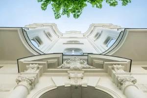 Blick von unten auf eine historische Fassade eines Gebäudes mit detaillierten architektonischen Verzierungen und Säulen. Im oberen Bereich des Bildes sind grüne Baumblätter und ein blauer Himmel zu sehen.