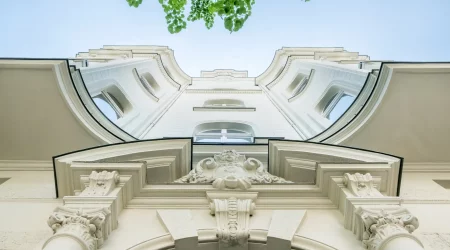 Blick von unten auf eine historische Fassade eines Gebäudes mit detaillierten architektonischen Verzierungen und Säulen. Im oberen Bereich des Bildes sind grüne Baumblätter und ein blauer Himmel zu sehen.