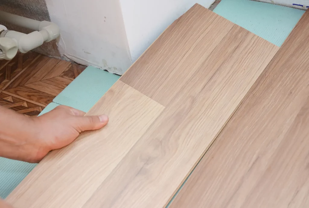 Handwerker verlegt Laminatboden, wobei die Paneele in einer bestimmten Richtung angeordnet werden. Nahaufnahme zeigt die korrekte Platzierung auf einer grünen Unterlage neben einem alten Holzparkett.