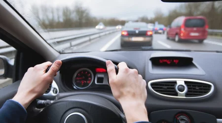 Ein Pendler fährt auf einer Autobahn zur Arbeit. Die Aufnahme zeigt die Perspektive des Fahrers mit beiden Händen am Lenkrad und Blick auf den Verkehr vor ihm