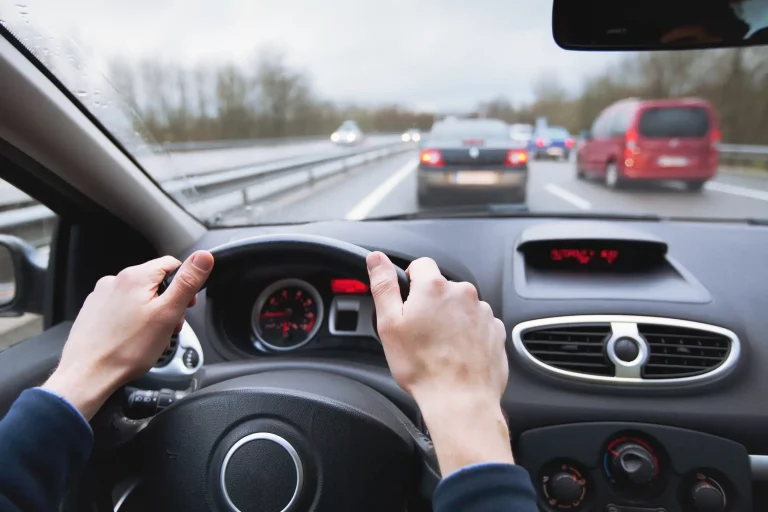 Ein Pendler fährt auf einer Autobahn zur Arbeit. Die Aufnahme zeigt die Perspektive des Fahrers mit beiden Händen am Lenkrad und Blick auf den Verkehr vor ihm