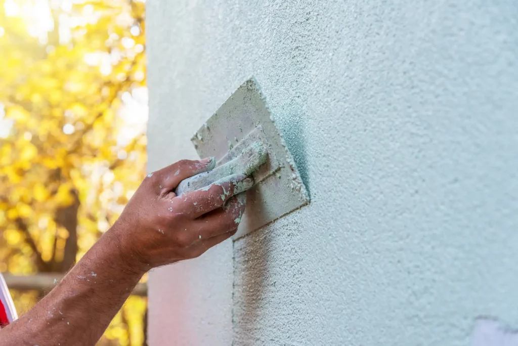 Handwerker, der mit einer Kelle eine Schicht Putz auf eine Außenwand aufträgt. Nahaufnahme zeigt die Technik und das Texturmuster des Putzes. Hintergrund unscharf mit gelb gefärbten Bäumen im Sonnenlicht.