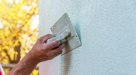 Handwerker, der mit einer Kelle eine Schicht Putz auf eine Außenwand aufträgt. Nahaufnahme zeigt die Technik und das Texturmuster des Putzes. Hintergrund unscharf mit gelb gefärbten Bäumen im Sonnenlicht.