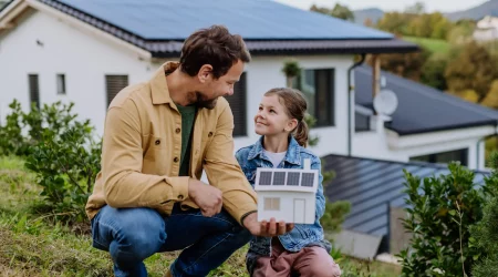 Ein Mann und ein Mädchen knien im Garten vor einem modernen Haus mit Solarpaneelen auf dem Dach. Das Mädchen hält ein Modellhaus mit Solarpaneelen, während sie und der Mann sich lächelnd ansehen.