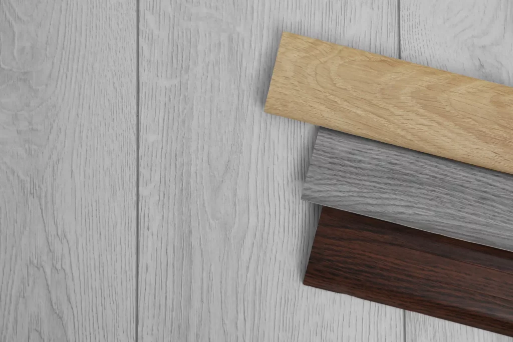 Verschiedene Laminat-Sockelleisten in unterschiedlichen Farben und Holzmustern auf einer hellgrauen Holzoberfläche angeordnet. Zeigt eine Auswahl von hellen, grauen und dunklen Holzdekoren, ideal zur Ergänzung von Laminatböden.