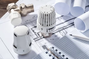Mehrere Thermostatventile liegen auf technischen Zeichnungen und Bauplänen. Daneben befinden sich aufgerollte Papierpläne, ein kariertes Notizbuch und ein Kugelschreiber. Die Anordnung dient der Heizungsberechnung für die Planung einer Heizungsanlage.