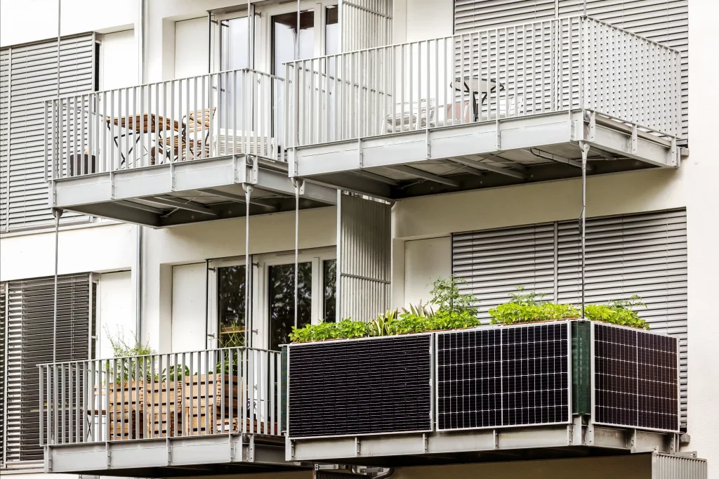 Balkonkraftwerk an einem modernen Wohngebäude. Auf dem unteren Balkon ist eine Solaranlage installiert, die Sonnenenergie nutzt. Der Balkon ist mit Pflanzen dekoriert, während die oberen Balkone Gartenmöbel aufweisen.