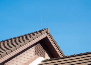 Blitzableiter auf dem Giebel eines Hauses mit braunem Ziegeldach unter klarem, blauem Himmel.