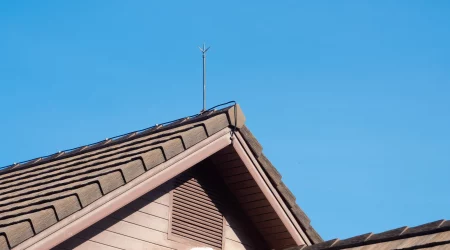 Blitzableiter auf dem Giebel eines Hauses mit braunem Ziegeldach unter klarem, blauem Himmel.