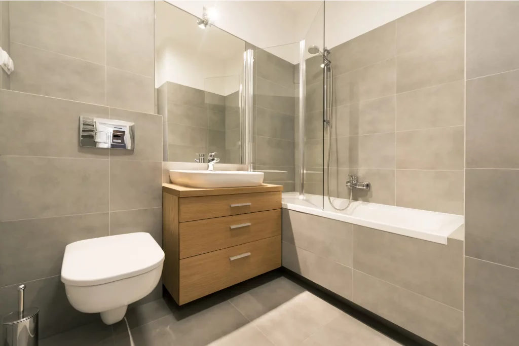 Modernes Badezimmer mit grauen Fliesen, einem hängenden WC, einem Waschbecken auf einem Holzunterschrank und einer Kombination aus Dusche und Badewanne. Die Dusche-Badewannen-Kombi hat eine Glaswand und eine verchromte Duscharmatur mit Handbrause.