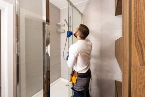 Ein Sanitärinstallateur in Arbeitskleidung und blauen Handschuhen installiert eine Duschwand einer neuen Dusche. Die teils geflieste Dusche hat eine eine weiße Duschtasse und modernisierte Armatur. Der Handwerker trägt Werkzeuge an einem Gürtel um die Hüfte.