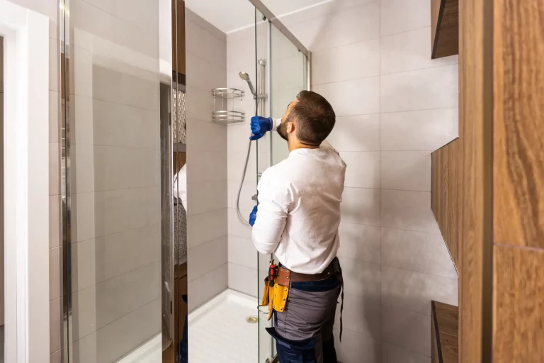 Ein Sanitärinstallateur in Arbeitskleidung und blauen Handschuhen installiert eine Duschwand einer neuen Dusche. Die teils geflieste Dusche hat eine eine weiße Duschtasse und modernisierte Armatur. Der Handwerker trägt Werkzeuge an einem Gürtel um die Hüfte.