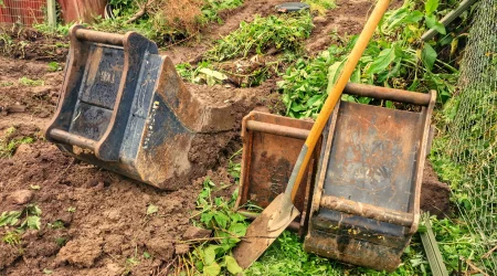 Spaten und Baggerschaufeln auf einer Gartenbaustelle mit frisch ausgehobenem Erdaushub, umgeben von Erde und Pflanzen