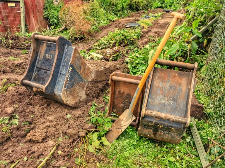 Spaten und Baggerschaufeln auf einer Gartenbaustelle mit frisch ausgehobenem Erdaushub, umgeben von Erde und Pflanzen