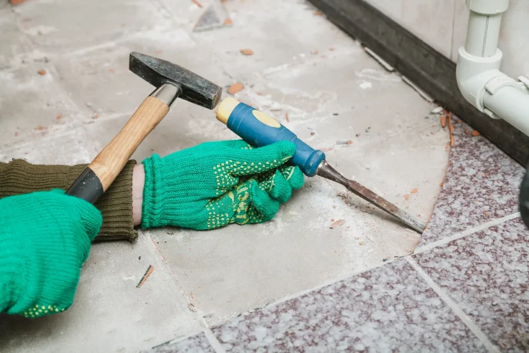 Ein Fliesenleger entfernt mit einem kleinen Meisel und einem Hammer verlegte Fliesen für die Vorbereitung einer Fußbodenheizung