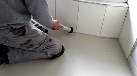Fliesenleger in Arbeitskleidung kniet auf dem Boden und trägt mit einer Kartuschenpistole frischen Dichtstoff auf die Fugen zwischen Bodenfliesen und der Wandverkleidung im Badezimmer auf.