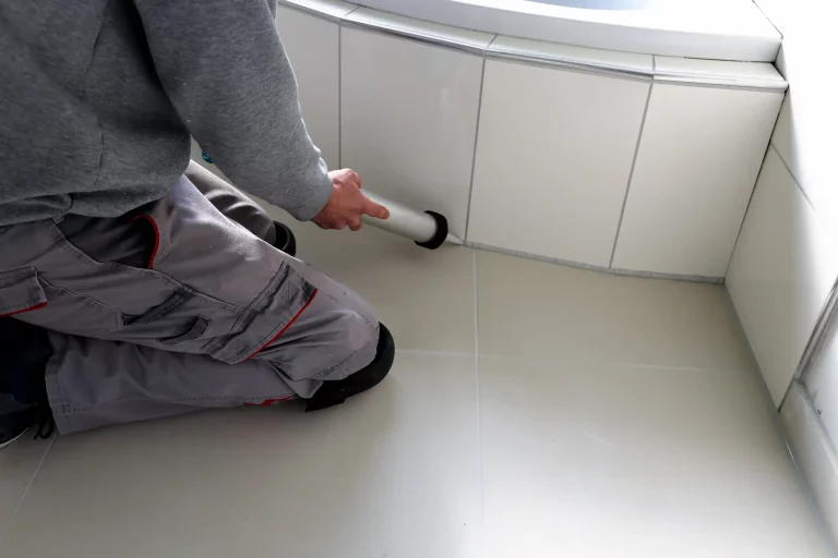 Fliesenleger in Arbeitskleidung kniet auf dem Boden und trägt mit einer Kartuschenpistole frischen Dichtstoff auf die Fugen zwischen Bodenfliesen und der Wandverkleidung im Badezimmer auf.