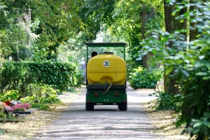 Ein kleines Fahrzeug eines Friedhofgärtners mit einem großen gelben Wassertank fährt auf einem schmalen Weg durch einen grünen, bewaldeten Friedhof. Der Tank hat eine große Zahl "20" auf der Rückseite. Der Weg ist von üppigem Grün und verschiedenen Pflanzen gesäumt, und im Vordergrund sind einige bunte Blumenbeete auf Grabstätten zu sehen.