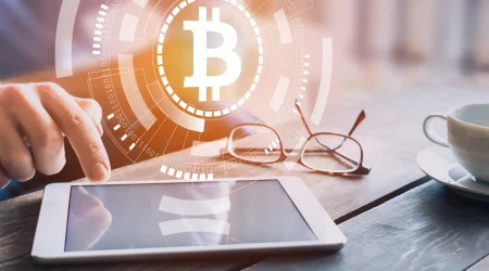 Person tippt auf ein Tablet auf einem Holztisch, im Hintergrund sind eine Brille und eine Tasse Kaffee. Im Vordergrund ist ein großes, leuchtendes Bitcoin-Symbol zu sehen, umgeben von technologischen und digitalen Symbolen.