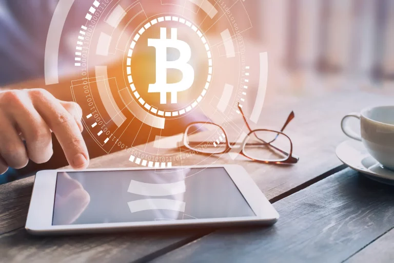 Person tippt auf ein Tablet auf einem Holztisch, im Hintergrund sind eine Brille und eine Tasse Kaffee. Im Vordergrund ist ein großes, leuchtendes Bitcoin-Symbol zu sehen, umgeben von technologischen und digitalen Symbolen.