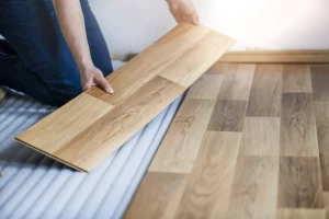 Nahaufnahme eines Handwerkers, der eine Laminatdiele verlegt. Das Bild zeigt, wie eine einzelne Laminatdiele passgenau auf den bereits verlegten Boden gesetzt wird.