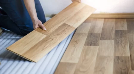 Nahaufnahme eines Handwerkers, der eine Laminatdiele verlegt. Das Bild zeigt, wie eine einzelne Laminatdiele passgenau auf den bereits verlegten Boden gesetzt wird.