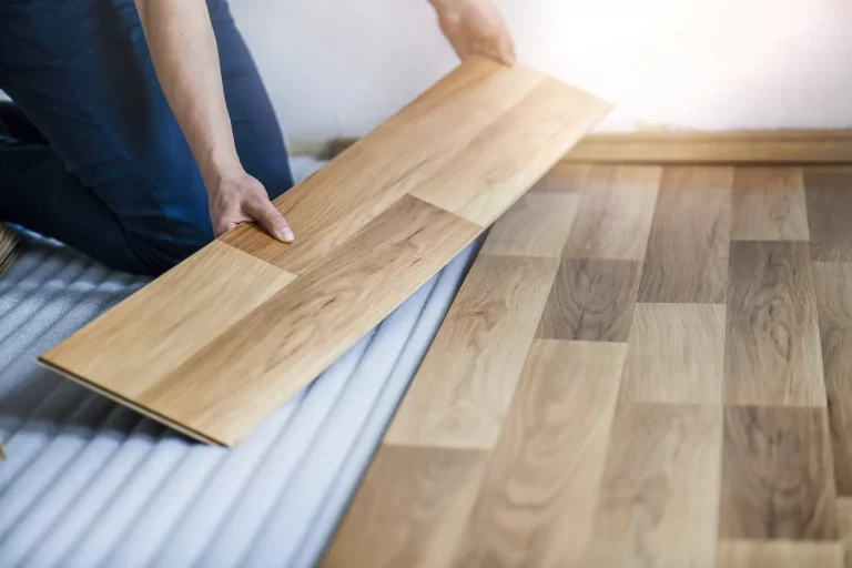Nahaufnahme eines Handwerkers, der eine Laminatdiele verlegt. Das Bild zeigt, wie eine einzelne Laminatdiele passgenau auf den bereits verlegten Boden gesetzt wird.