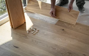 Handwerker verlegt Laminatboden in einem Raum mit großen Fenstern. Eine Laminatdiele wird in die bereits verklebte Fläche eingesetzt, während mehrere weitere Dielen und Abstandshalter auf dem Boden liegen.