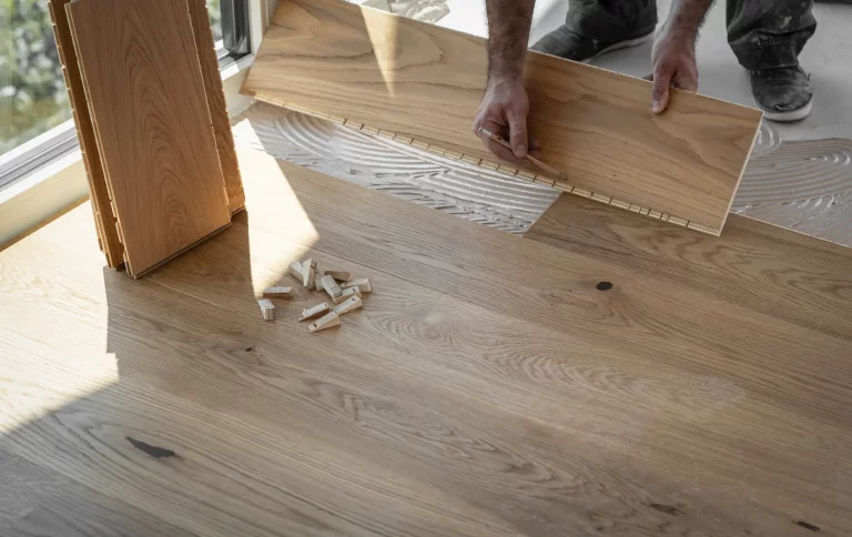 Handwerker verlegt Laminatboden in einem Raum mit großen Fenstern. Eine Laminatdiele wird in die bereits verklebte Fläche eingesetzt, während mehrere weitere Dielen und Abstandshalter auf dem Boden liegen.
