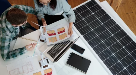 Zwei Personen arbeiten an der professionellen Planung einer Photovoltaikanlage, mit Solarpanelen, Gebäudeplänen und einem Laptop auf dem Tisch.