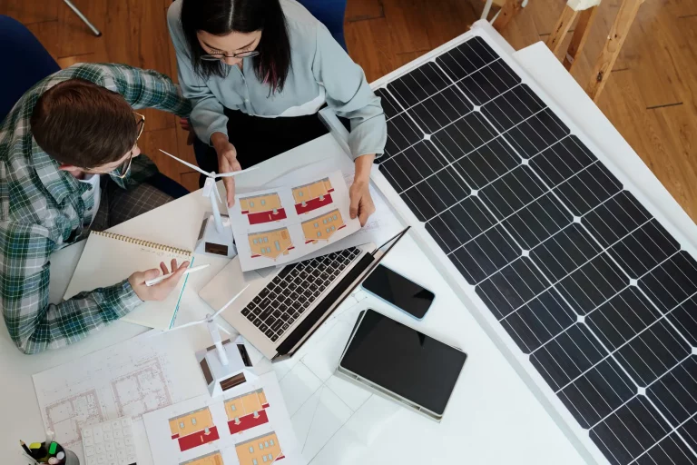 Zwei Personen arbeiten an der professionellen Planung einer Photovoltaikanlage, mit Solarpanelen, Gebäudeplänen und einem Laptop auf dem Tisch.