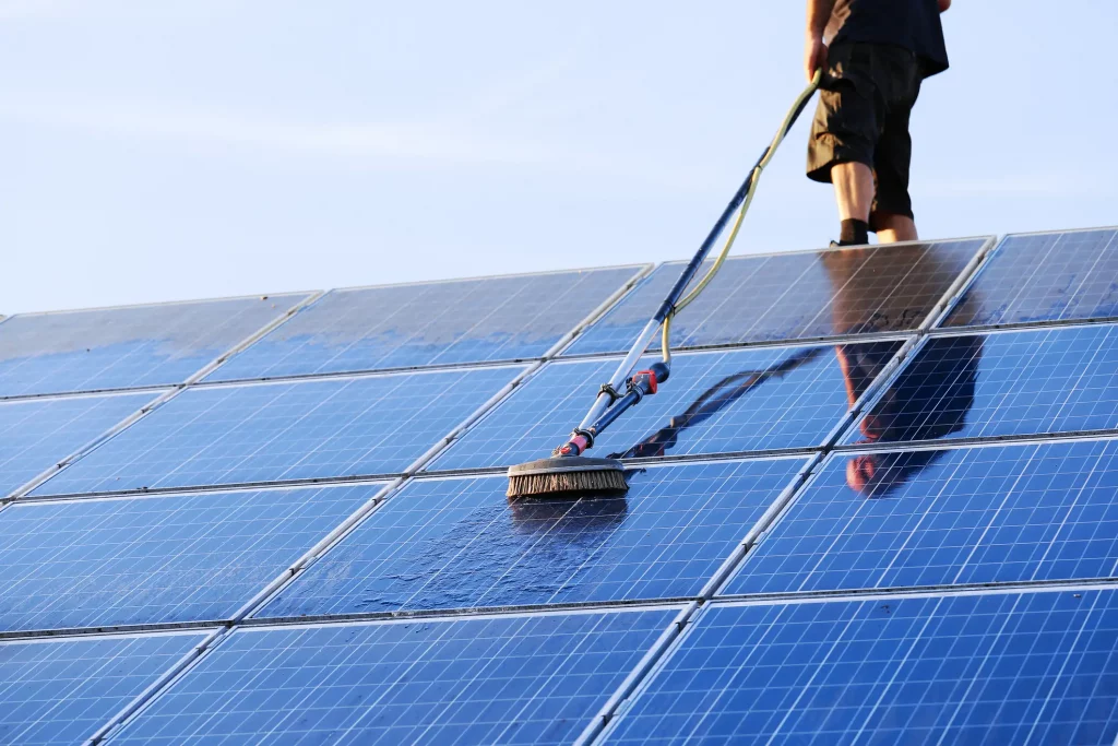 Person reinigt Solarmodule auf einem Dach mit einer Bürste und einem Wasserschlauch bei klarem Himmel.