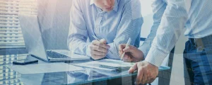 Ein Unternehmer und ein Steuerberater sitzen an einem Schreibtisch und besprechen die Steuerberatung für eine GmbH. Auf dem Tisch liegen ein Laptop, ein Smartphone und Dokumente, während beide konzentriert Notizen überprüfen und Papiere unterzeichnen.