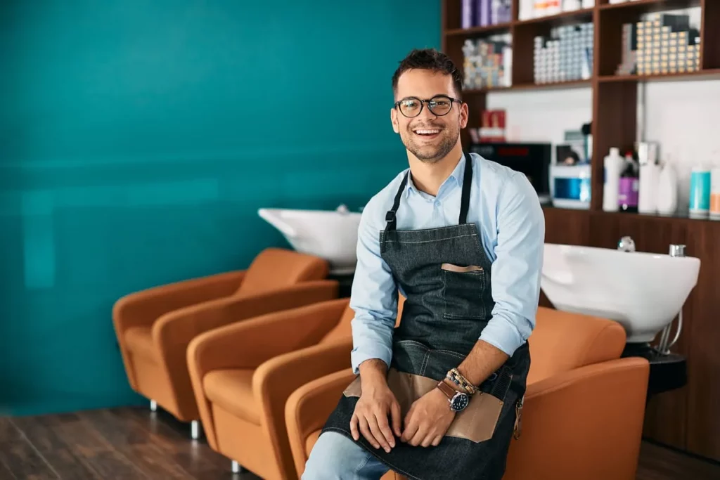 Ein lächelnder Friseur mit Brille und Schürze sitzt in einem modernen Friseursalon. Im Hintergrund sind Friseurstühle und Waschbecken zu sehen, sowie Regale mit Haarpflegeprodukten.