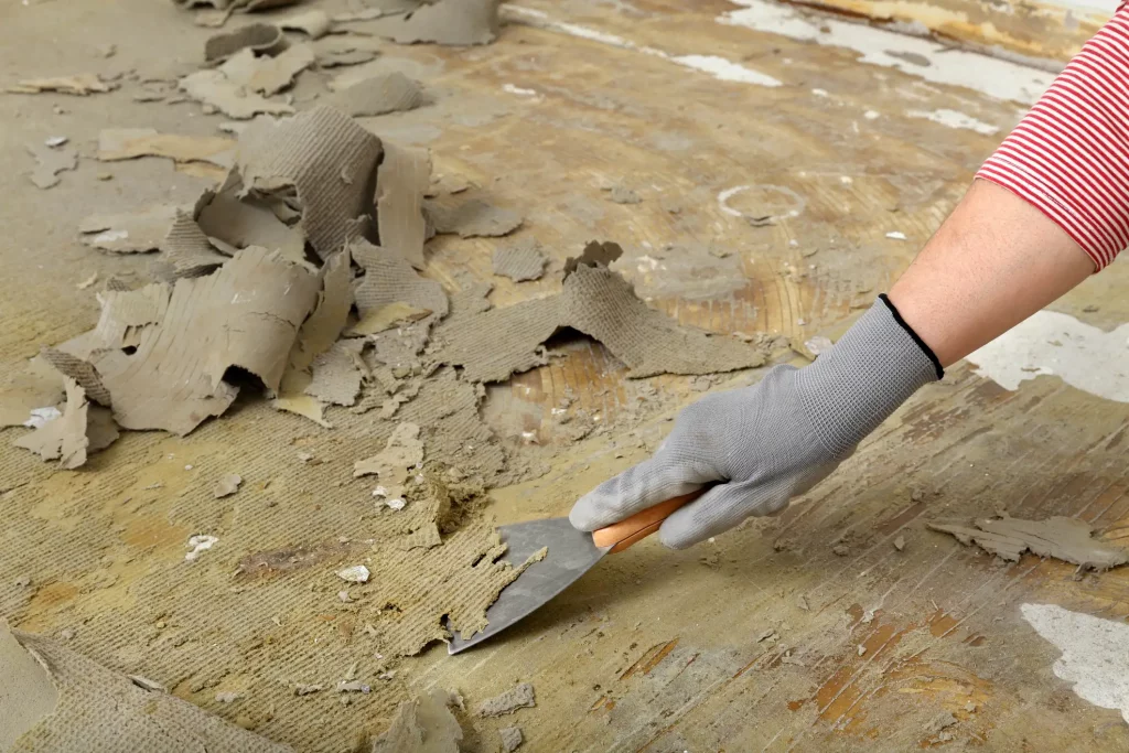 Eine Hand, die einen Spachtel benutzt, um alte Klebereste eines Teppichbodens zu entfernen. Die Person trägt einen grauen Handschuh und ein rot-weiß gestreiftes Hemd. Der Boden ist teilweise abgedeckt mit abblätternden Schichten des alten Teppichklebers.