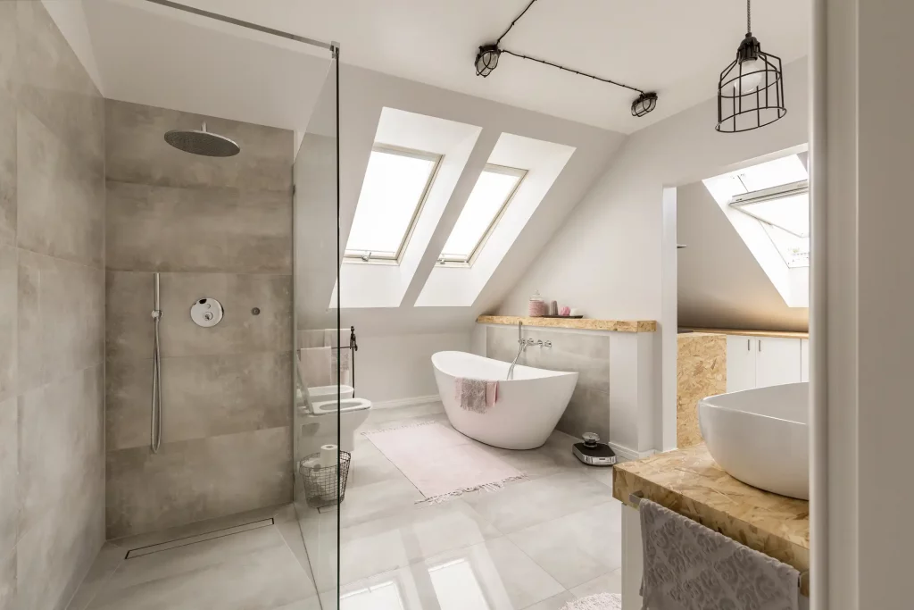 Modernes Badezimmer mit Dachfenstern, einer freistehenden Badewanne und einer bodengleichen Walk-In-Dusche. Die Dusche hat eine Glaswand, eine Regenbrause und eine Handbrause. Die Wände der Dusche sind mit beigen Fliesen verkleidet. Im Raum befinden sich zudem eine freistehende weiße Badewanne, ein Holzregal über der Wanne und rosa Akzente wie Handtücher und Teppiche.
