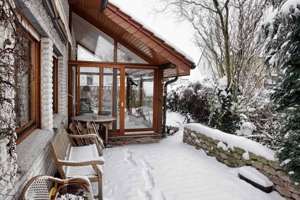 Warmwintergarten mit großen Holzfenstern und einer verglasten Schiebetür, die zu einer schneebedeckten Terrasse führt. Der Außenbereich ist von Schnee bedeckt, einschließlich der Holzbänke und des Tisches. Karge Bäume und Sträucher umgeben den Wintergarten und verstärken die winterliche Atmosphäre.
