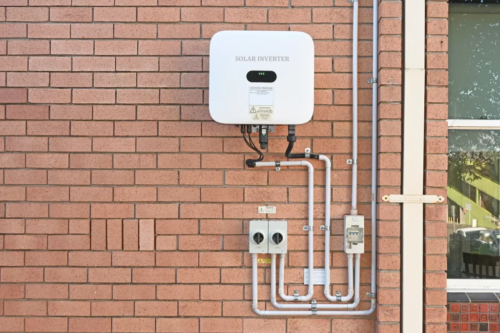 Ein Wechselrichter für Photovoltaikanlagen ist an einer Ziegelwand montiert. Unter dem Wechselrichter verlaufen mehrere Rohre und Kabel zu zwei Schaltern, die ebenfalls an der Wand befestigt sind. Rechts neben der Installation befindet sich ein Fenster.