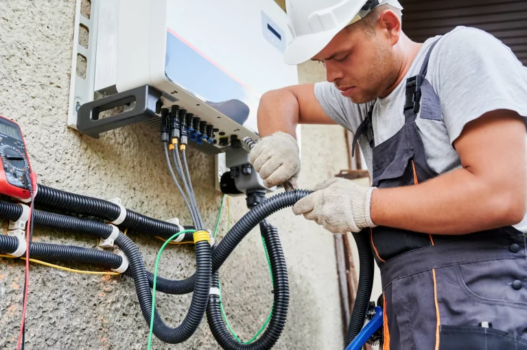 Ein Elektriker in Arbeitskleidung und Schutzhelm schließt einen PV-Anlagen Wechselrichter an einer Wand an. Der Wechselrichter ist mit mehreren Kabeln verbunden, die in schwarzen Kabelschutzschläuchen gebündelt sind. Auf der linken Seite ist ein Multimeter an den Kabeln angebracht, um elektrische Messungen durchzuführen.